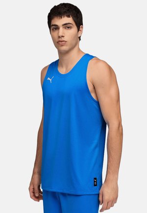 Mladý muž ve světlém modrém sportovním tílku Puma a odpovídajících šortkách stojí na jednoduchém světlém pozadí.