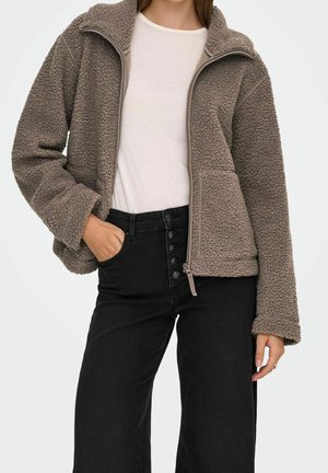 Kobieta w taupe polarowej kurtce na zamek, nosząca ją na białej koszuli oraz czarne, wysokowalne dżinsy z guzikiem z przodu, stoi z jedną ręką w kieszeni.