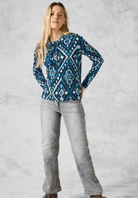 Lange mouw hoodie in het blauw met geometrisch patroon, gecombineerd met vervaagde grijze jeans. De stof lijkt lichtgewicht te zijn met een aansluitende pasvorm.