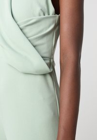 Combinaison sans manches vert menthe avec un détail de tissu drapé à la taille et une texture lisse, complétée par un bras tonique à côté.