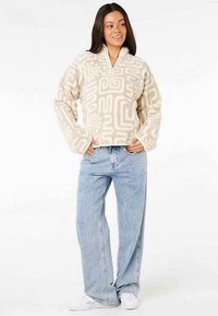 Kremowy polar z abstrakcyjnymi beżowymi wzorami, z dekoltem na zamek do ćwiartki, szerokimi rękawami, noszony z jasnoniebieskimi jeansami o szerokich nogawkach.