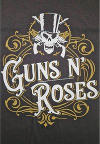 Černé tričko s bílým a zlatým grafickým designem, zahrnujícím lebku s cylindrem, pistole a výrazně zobrazený text "GUNS N' ROSES".