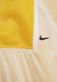 Nike-topp med en gul nätpanel, slät krämfärgad tyg, kontrasterande svart logotyp. Lätt och ventilerande design med distinkta texturer.