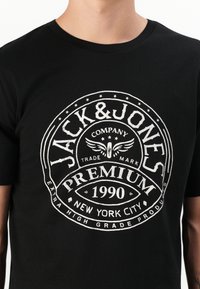 Svart t-skjorte laget av bomull, med et stort hvitt grafisk trykk av "Jack & Jones Premium 1990" og dekorative stjerner og vinger.