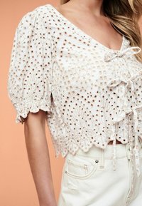 Hvid cropped bluse med indviklet broderi, korte pufærmer, v-hals og bindebånd foran med detaljerede bølgede kanter.