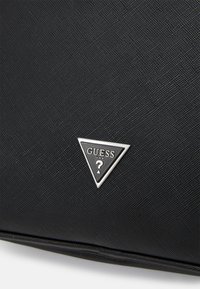 Sac en tissu noir texturé avec un logo matériel triangulaire argenté portant "GUESS" et un point d'interrogation. Motif en relief subtil sur la surface.