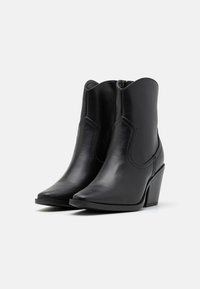 Botas de cuero negro hasta el tobillo con punta afilada y tacón en ángulo. Textura suave con costuras decorativas y una abertura curva en la parte superior.