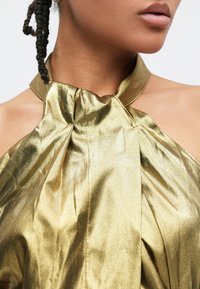 Haut à col licou en tissu métallisé doré plissé, avec un détail nœud au niveau de l'encolure et une texture lisse et brillante.