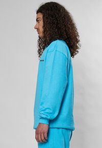Lyseblå genser med lange ermer, rund hals, og brodert tekst "SWIM SE." Myk tekstur, avslappet passform, sidevisning.