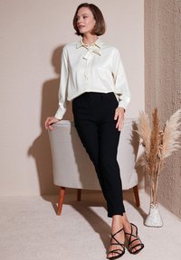 Blusa de satén crema con detalle de lazo, combinada con pantalones negros de cintura alta. La modelo está de pie al lado de una silla de color claro y un jarrón decorativo.