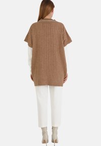 Brązowy prążkowany, oversize sweter z dzianiny z kołnierzykiem, krótkimi rękawami i rozcięciami po bokach, zestawiony z białymi spodniami i beżowymi botkami na obcasie.