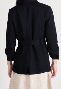 Žena v černém s páskem nad béžovou sukni, zobrazená zezadu na jednobarevném světlém pozadí.