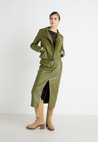 Veste de moto en simili cuir vert olive et jupe midi assortie avec une fente, associées à des bottes en daim beige montantes.