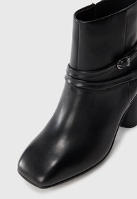 Black leather ankle boot with square toe, block heel, and decorative strap with silver buckle on white background.