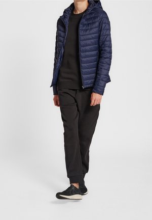 Person iført en marineblå quiltet hættetrøje, sort sweatshirt, sorte joggingbukser og sorte træningssko, stående mod en ensfarvet baggrund.