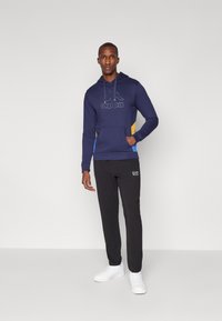 Hoodie azul-marinho com um logo, painéis laterais amarelos e azuis em contraste, e bolso frontal; combinado com calças pretas e sapatos brancos.