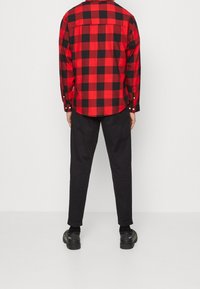 Camisa de cuadros rojos y negros de manga larga con puños abotonados, combinada con pantalones negros y zapatos negros, vista desde atrás.