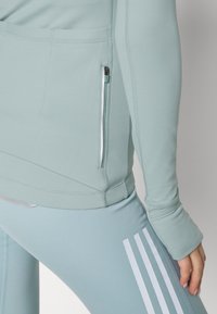 Personne portant une tenue de sport bleu clair avec une poche zippée sur le côté de la veste et trois bandes blanches sur les leggings.
