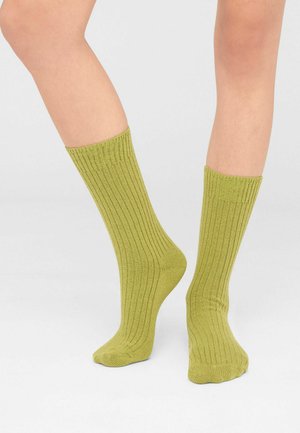 Una modelo lleva calcetines acanalados de color verde oliva que llegan a media pantorrilla y levanta ligeramente un pie sobre un fondo blanco.