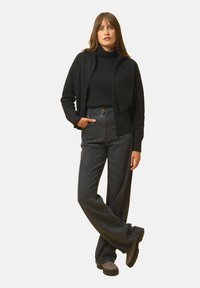 Musta vetoketjullinen neuletakki, korkeakauluksinen paita ja korkeavyötäröiset tummat farkut. Mallilla harmaata platomainen kengät, seisoo yhdellä kädellä lanteellaan.