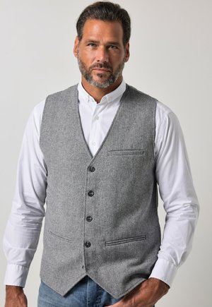 Mand med kort mørkt hår og skæg iført en grå knapvest over en hvid skjorte og blå jeans, stående mod en ensfarvet baggrund.