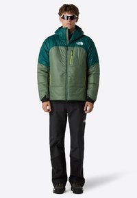 Heren pufferjack in groen en teal met een ritszak, voorzien van zwarte accenten en een capuchon, gecombineerd met zwarte waterdichte broeken.
