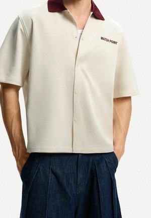 Koszula kremowa z krótkim rękawem, bordowym kołnierzykiem i napisem "MATCH POINT", noszona na wierzchu ciemnoniebieskich plisowanych spodni.