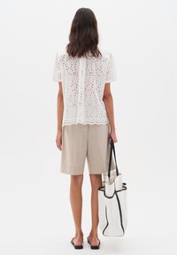 Camicetta bianca a maniche corte con dettagli in pizzo, abbinata a pantaloni corti in lino beige. Una borsa a tracolla bianca con accenti neri è tenuta in mano.