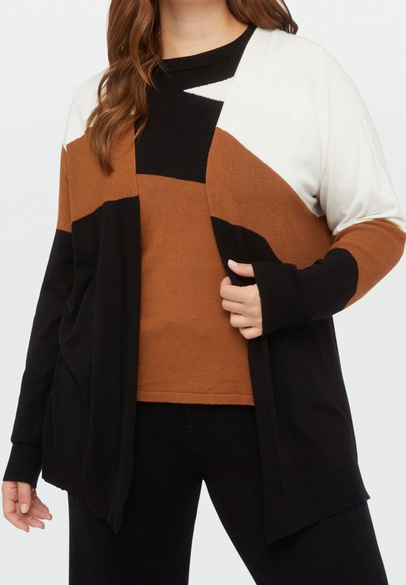 Donna che indossa un cardigan a blocchi di colore bianco, marrone e nero su un top in maglia abbinato marrone e nero, abbinato a pantaloni neri.