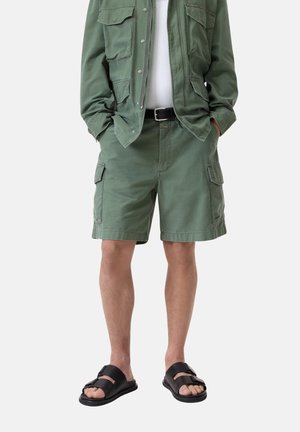 Pantaloni cargo in cotone verde oliva, con molteplici tasche, abbinati a una cintura nera e sandali neri scivolabili.