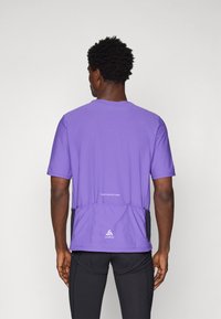 Camiseta deportiva morada de mangas cortas, con dobladillo curvado, bolsillos traseros y texto reflectante. Confeccionada con un tejido transpirable y de textura suave.