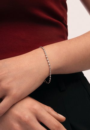 Silberfarbener Armband mit einer Reihe kleiner, länglicher Perlen, die am Handgelenk getragen wird. Im Hintergrund ist ein tiefrotes Oberteil und eine dunkle Hose zu sehen.