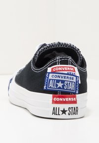 Svart canvas Converse-skor har vita gummisulor och flera märkespatchar i röd och blå vid hälen, med kontrasterande vit söm.