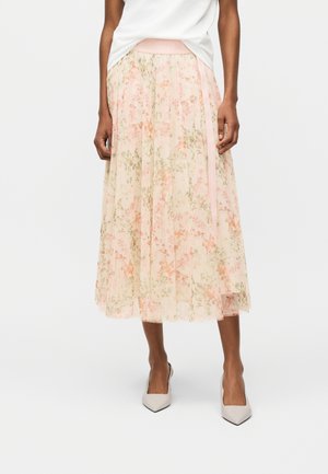 Frau trägt einen hellrosa Midirock aus floralem Tüll mit einem satinband an der Taille, ein weißes Oberteil und beige spitze High Heels.