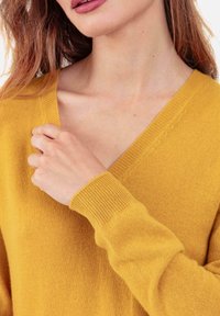 Pull en maille jaune moutarde avec un col en V et des poignets côtelés. Texture douce, coupe ajustée, avec de légers détails de surpiqûres le long du col.