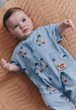 Baby, das auf einer braunen gesteppte Decke liegt und einen blauen Strampler mit Hasenmotiven und einem Patch mit dem Schriftzug „Hello Fun“ trägt.
