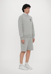 Hanorac gri cu manșete și tiv din țesătură cu nervuri, având un logo negru; purtat cu pantaloni scurți gri asortati și adidași albi.