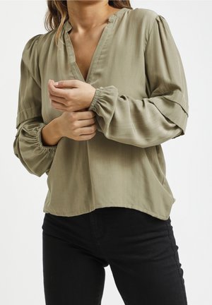 Frau trägt eine olivgrüne Bluse mit langen Ärmeln und elastischen Bündchen sowie schwarze Hosen und richtet mit der rechten Hand den linken Ärmel.