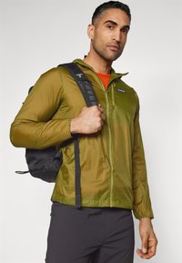 Green lightweight jacket with a full zipper, textured fabric, long sleeves, and a hood, paired with a black backpack against a light background.