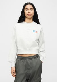 Γυναίκα με μακριά σκουρόχρωμα μαλλιά που φοράει ανοιχτό γκρι cropped φούτερ με μπλε λογότυπο και σκούρο γκρι παντελόνι με ελαστική μέση, στέκεται μπροστά σε λευκό φόντο.