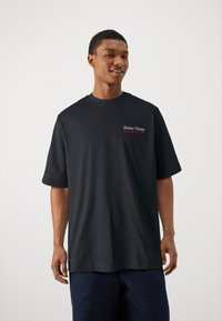 Sort bomulds t-shirt med korte ærmer, rund halsudskæring og et lille broderet mærke i rød og hvid på brystet. Glat stofstruktur.