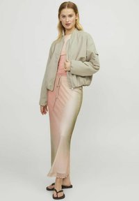 Beige bomberjack over een roze-groene ombre jurk; de jurk heeft een trekkoord in de taille en een zachte satijnen textuur met een rafelige zoom. Zwarte slippers.