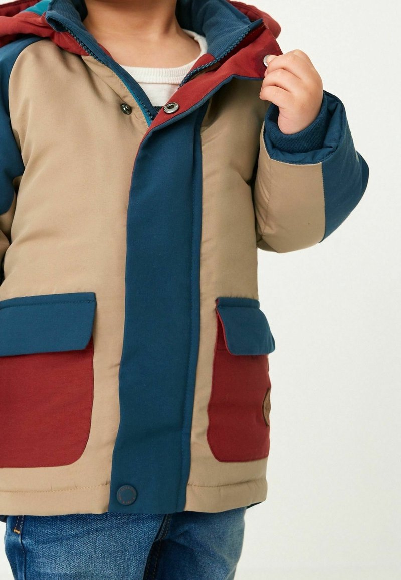 Enfant portant une veste matelassée color-block beige, bleue et rouge avec des poches, tenant le col de la veste d'une main.