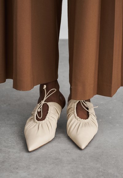 Chaussures en cuir crème à bout pointu avec un design froncé, des détails à lacets et une découpe à l'avant, assorties à un pantalon large marron.