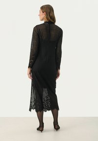 Langes schwarzes Kleid aus gemustertem Stoff, mit durchsichtigen Abschnitten und Spitzenverzierung am Saum. Hat lange Ärmel und einen hohen Halsauschnitt.