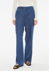 Jeans ampia blu in denim con una consistenza liscia, caratterizzati da un'alta vita e delicate rifiniture a cucitura. I pantaloni sono abbinati a slip-on floreali.