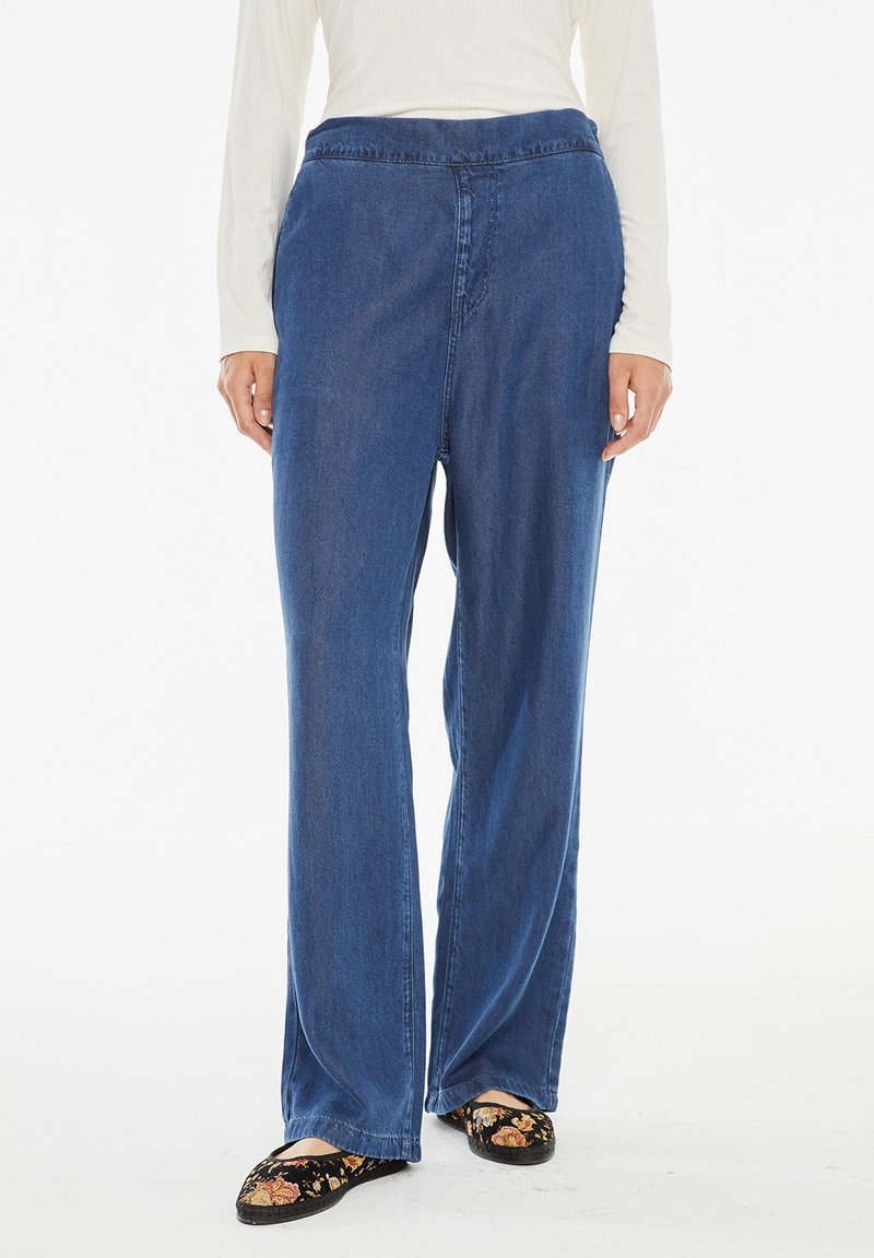 Jeans ampia blu in denim con una consistenza liscia, caratterizzati da un'alta vita e delicate rifiniture a cucitura. I pantaloni sono abbinati a slip-on floreali.