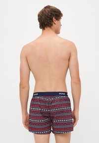 Costume de baie cu model în nuanțe de bleumarin, roșu și alb, având un design repetat cu "HUGO", talie elastică și o potrivire confortabilă.