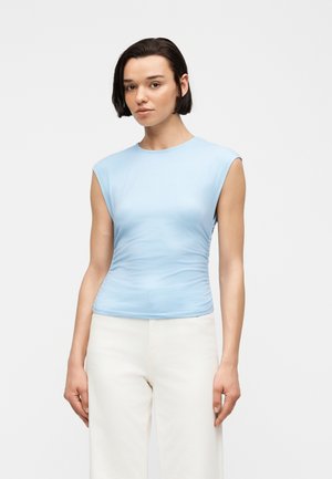 Jeune personne aux cheveux courts et foncés portant un haut sans manches bleu clair et un pantalon blanc, debout devant un fond uni.