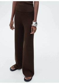 Pantalons en maille marron à jambes larges avec taille haute, associés à des tongs noires. Un bracelet tordu en argent orne le poignet.